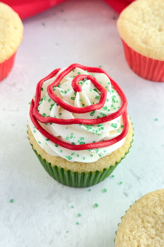 A vanilla cupcake frosted with vanilla buttercream frosting and decorated with green sanding sugar and red buttercream stripes.