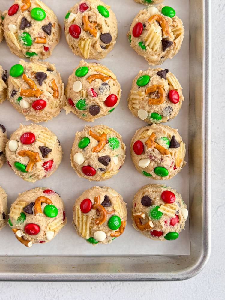 Cookie dough balls on a baking pan with additional chocolate chips, M&Ms, pretzels, and potato chips pressed in.