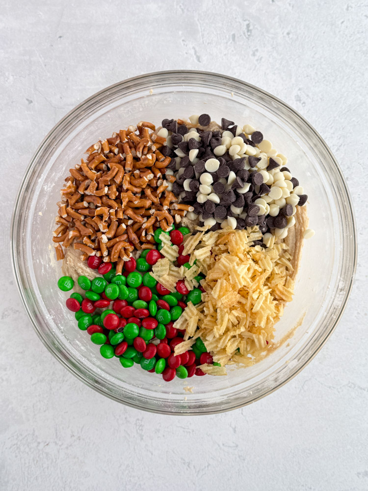 White and semisweet chocolate chip, holiday M&Ms, potato chips, and pretzels in a mixing bowl with cookie dough.
