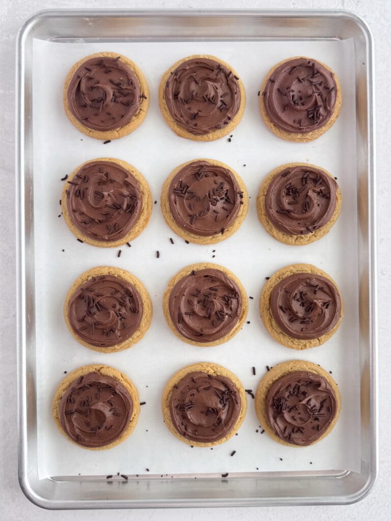 Chocolate buttercream frosted peanut butter cookies with chocolate sprinkles on a baking sheet pan.