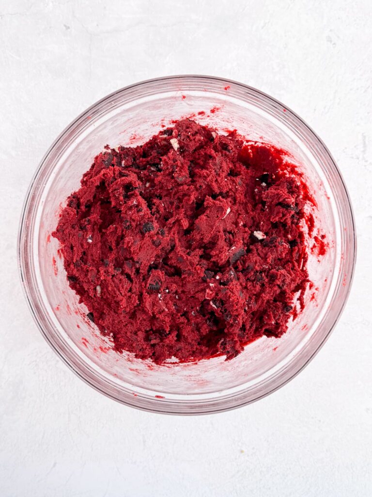 Red velvet cookie dough with chocolate chips and Oreo cookie pieces in a glass mixing bowl.