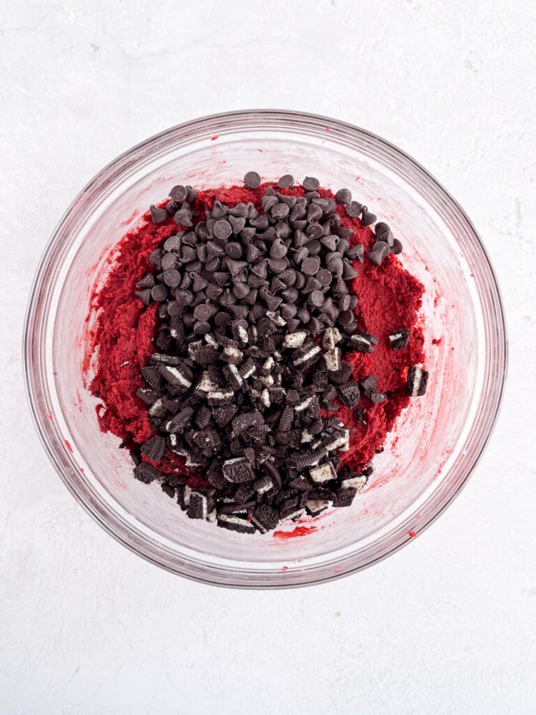 Red velvet cookie dough with chocolate chips and Oreo cookie pieces in a glass mixing bowl.