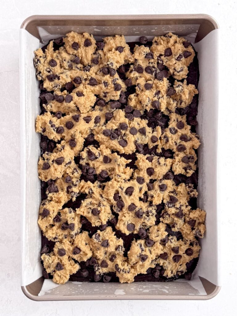 Chocolate chip cookie dough on brownie batter in a baking pan with additional chocolate chips.