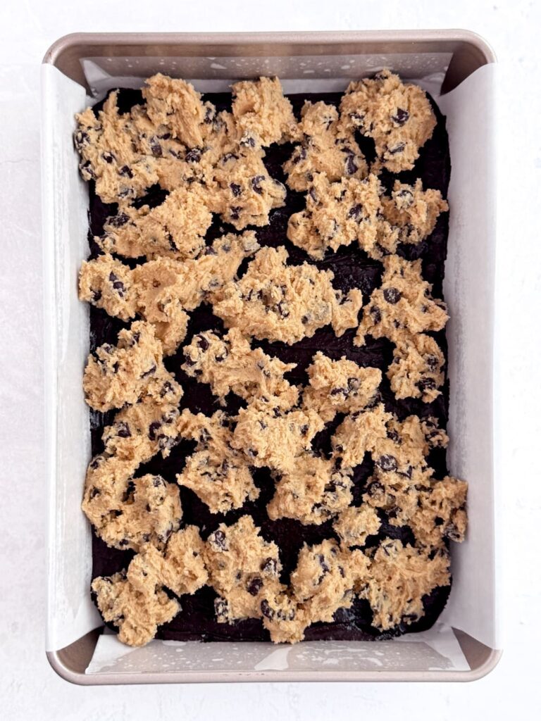 Chocolate chip cookie dough on brownie batter in a baking pan.