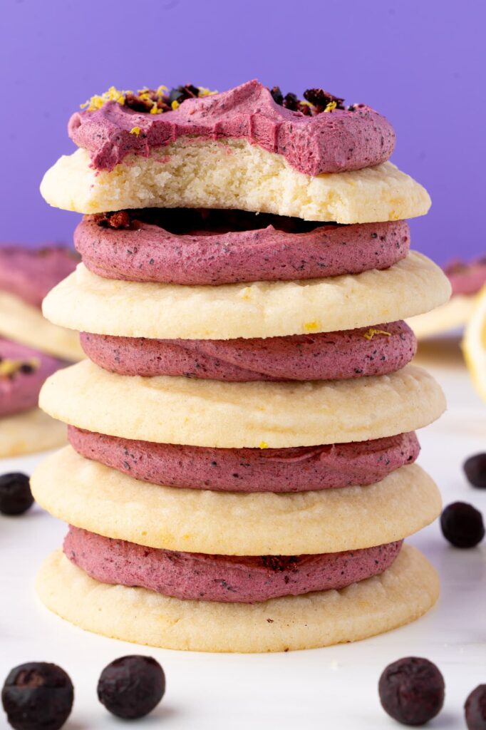 A stack of lemon cookies with blueberry frosting with a bite taken out of the top cookie.