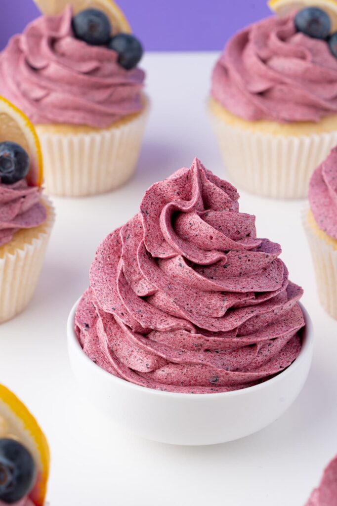 A swirl of blueberry frosting in a small white bowl with cupcakes in the background.