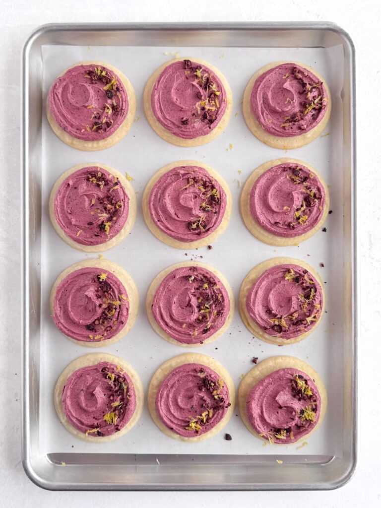 Lemon cookies frosted with blueberry buttercream and garnished with freeze-dried blueberries and lemon zest on a baking sheet pan.