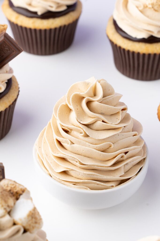 A swirl of toasted marshmallow frosting in a small white bowl with s'mores cupcakes in the background.