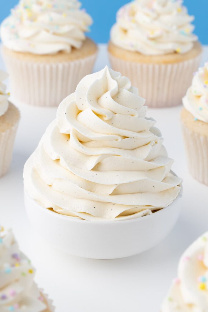 A swirl of vanilla buttercream frosting in a small white bowl with cupcakes in the background.