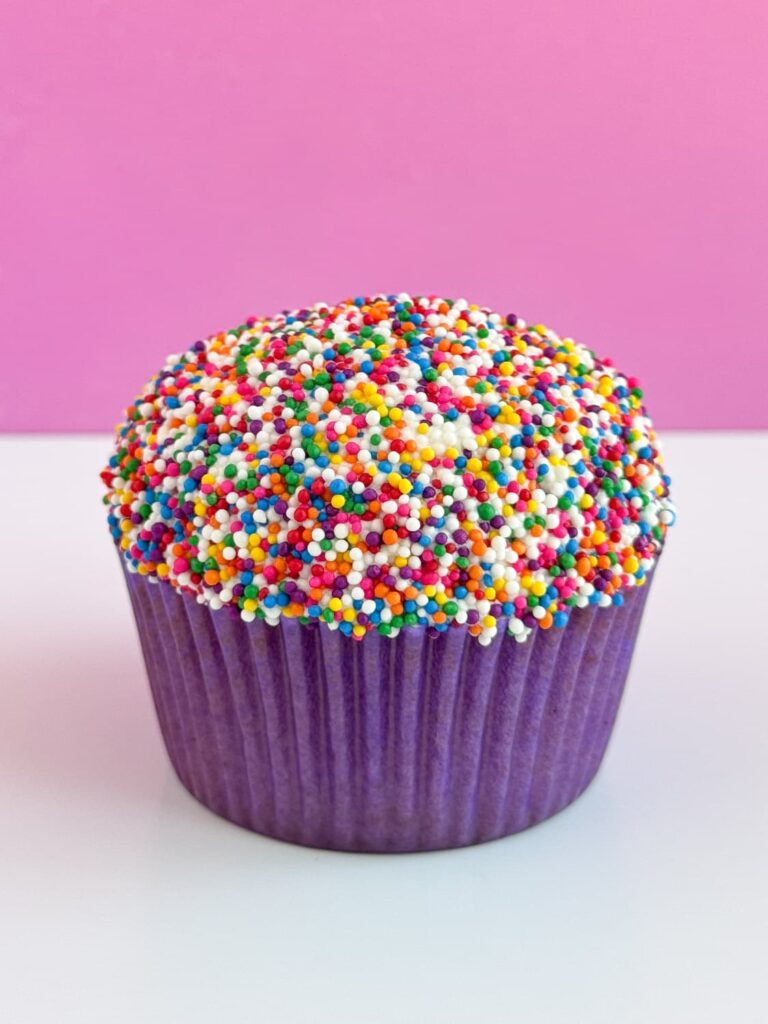 Vanilla cupcake in a purple cupcake liner with vanilla frosting and rainbow nonpareil sprinkles.