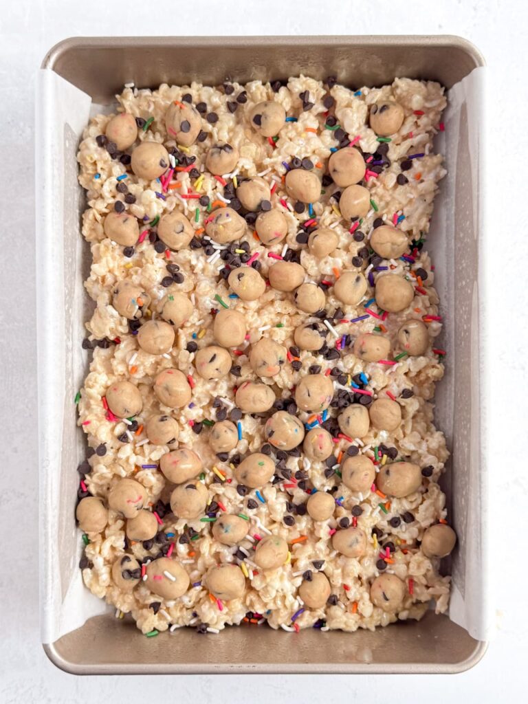 The Rice Krispie treat mixture pressed into a baking sheet pan with cookie dough pieces, chocolate chips, and rainbow sprinkles on top.
