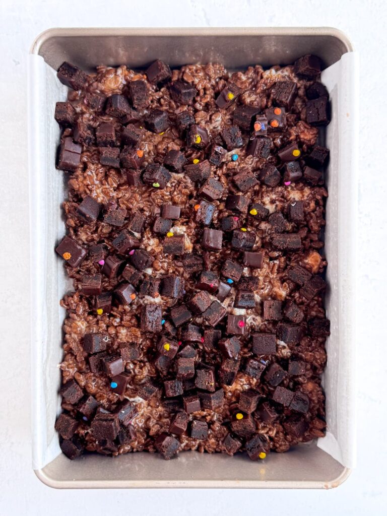 Chocolate Rice Krispie treat mixture spread into a baking pan with cosmic brownie pieces on top.