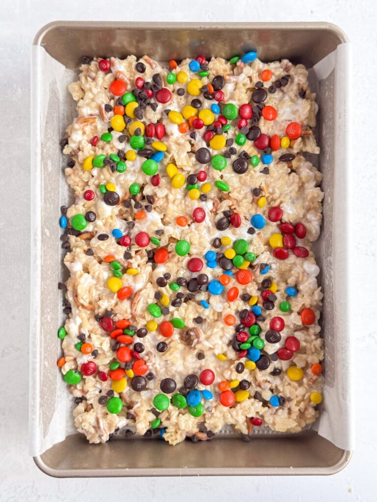 M&Ms and chocolate chips sprinkled onto Rice Krispie treat mixture in a baking pan.