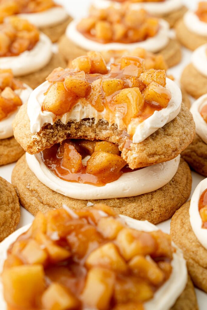 A bite taken out of a frosted caramel apple cookie showing the soft, chewy cinnamon brown sugar cookie beneath salted caramel buttercream, cinnamon apples, and caramel sauce.